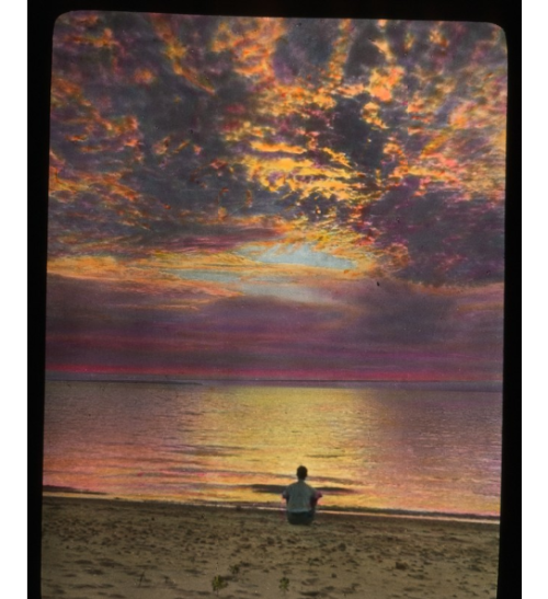 Hand tinted Lantern slide showing an image of a man sitting on beach at sunset.Isle Royale, MI 1930