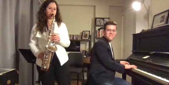Image: A woman, Alexa Tarantino, stands and plays a saxophone while a man, Steven Feifke, plays the piano. They are in a living room setting.