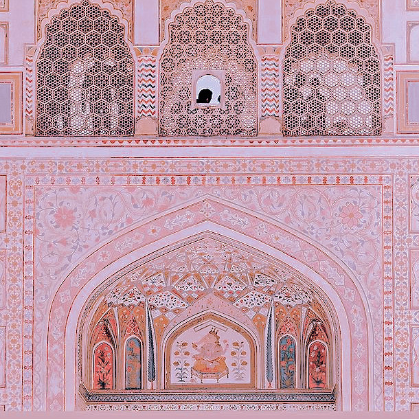 a front view of a bright pink house in rajasthan, with the traditional latticed "jaali" windows and elaborate arched doorways covered in mosaics.