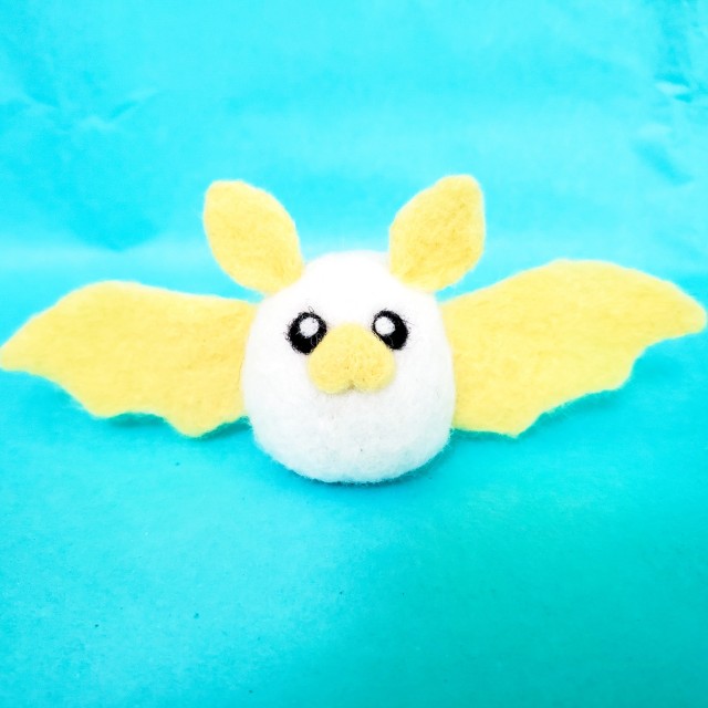 A photo of a white needle felted bat with a yellow nose, ears, and wings. It is sitting on a bright blue background