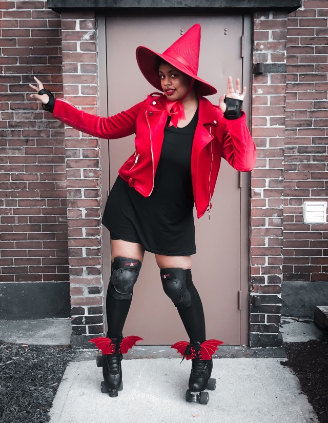 black person in a red witch hat, red moto jacket, black dress, roller skates with bat wing shoe ties, knee pads and wrist guards smiling while posing. 