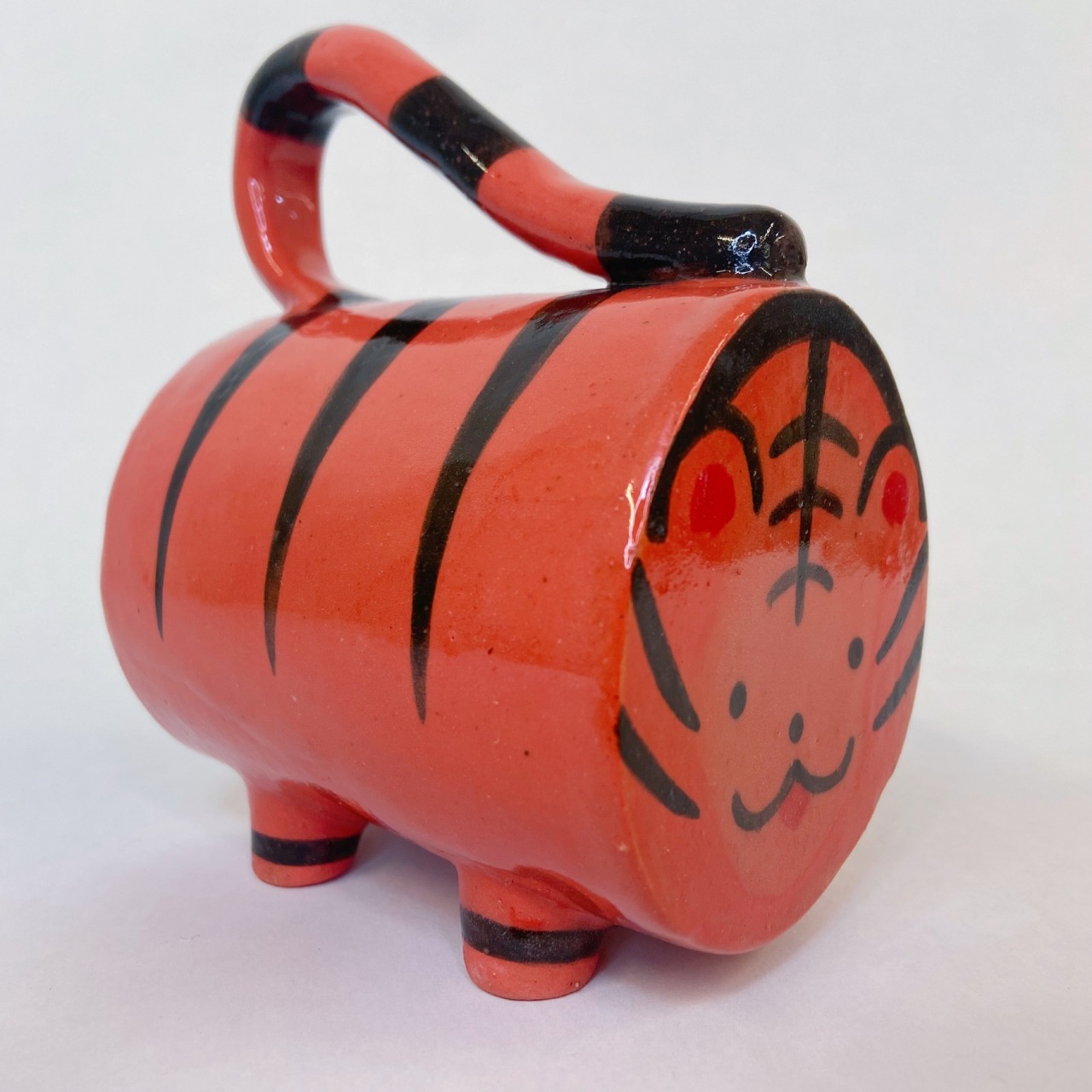 Photo of handmade ceramic tiger mug. It is red-orange with black stripes. The handle of the mug is the tail, and the bottom of the mug is the face. On the other side of where the handle is, there are four feet so the mug can stand up sideways.