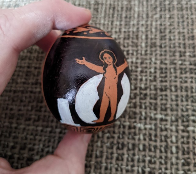 An egg that has been drawn on to resemble an ancient Greek red figure vase depicting Helen, small but fully formed, standing with her arms raised between two halves of an egg shell. This is from an angle to the left.