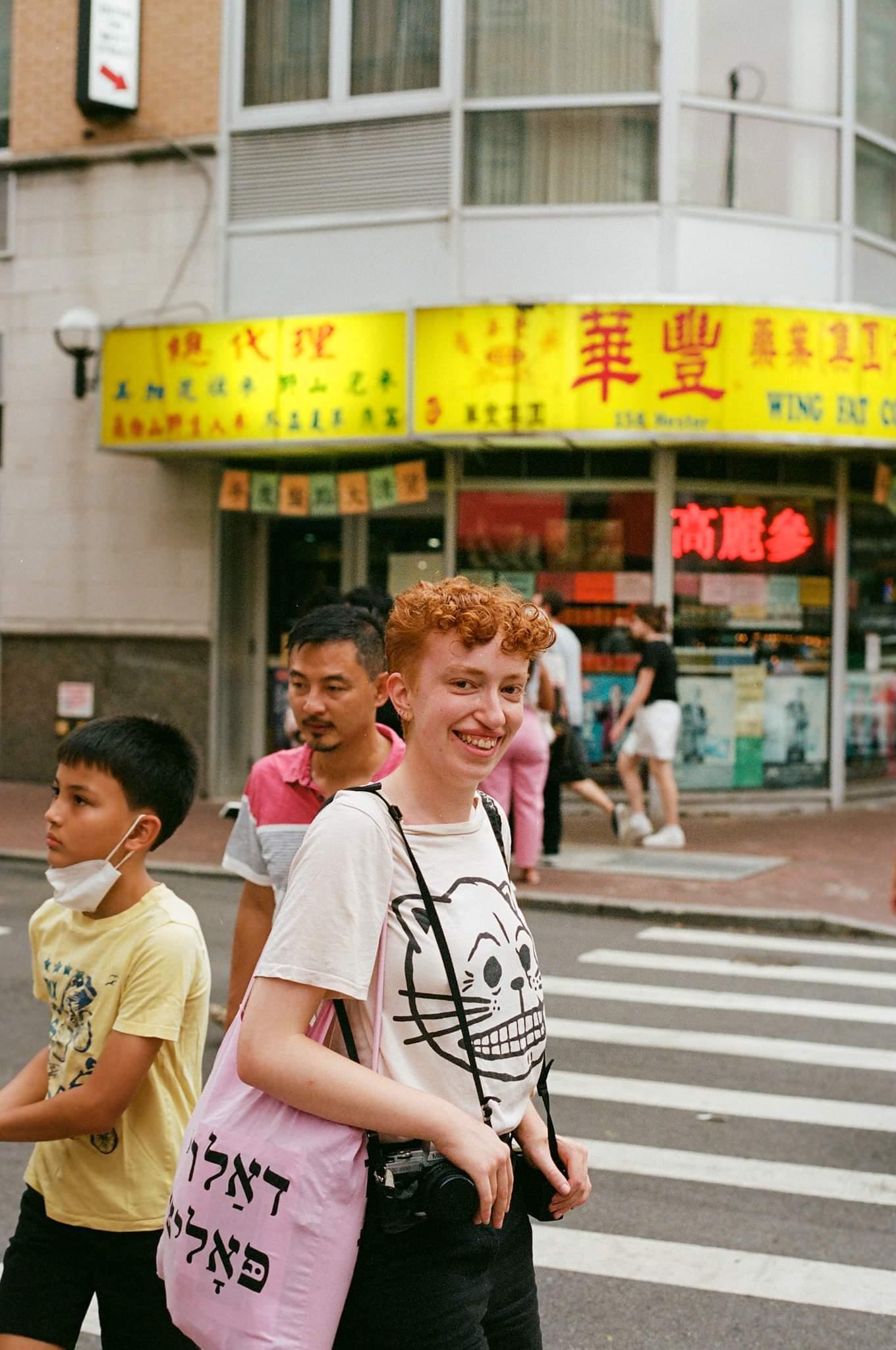 a film photograph of Storm, a white person with short ginger hair, smiling in front of a chinatown shopfront, holding 2 cameras. phot by Daniel Velez @djvelezc.
