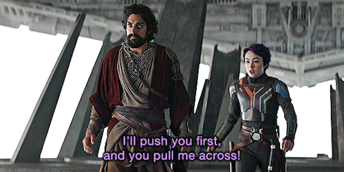 Gif from Ahsoka episode 1.08. Ezra stands on a platform staring out over a chasm as Sabine backs up behind him, saying, "I push you first, and you pull me across!" Ezra looks back at her.