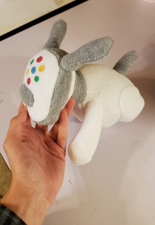 A photograph of a plushie made to resemble an iDog. This one is white and turned to the side, with multicolored spots on the face.