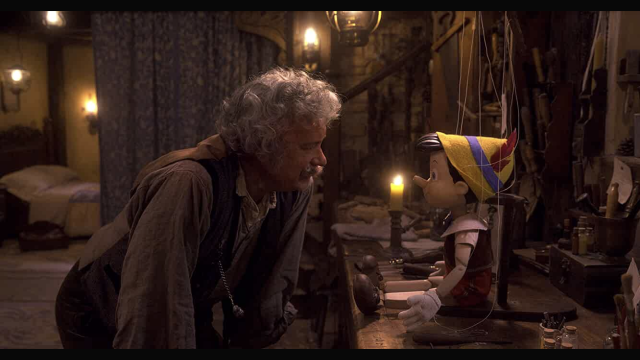 Tom Hanks as Geppetto with gray hair/beard bent to look at Pinocchio on his work bench