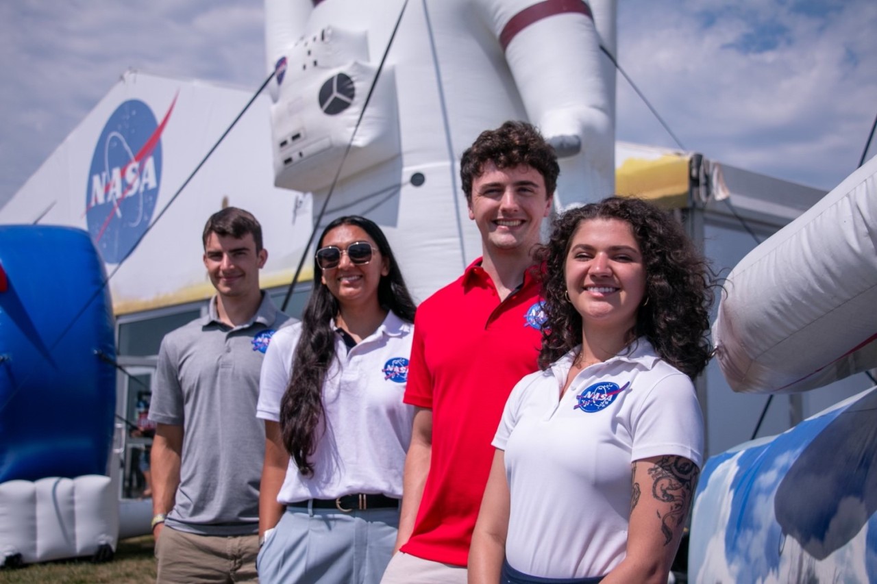 Four people pose in front of a giant inflatable astronaut. Each of the four people are in polos with a NASA insignia on the upper left of their shirt. It’s sunny and clouds and a blue sky can be seen in the background. A large white tent with a NASA logo emblazoned above the entrance can also be seen in the background. Credit: NASA
