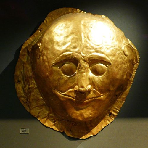 [image description: a mycenaean death mask made out of hammered gold. it has wide, pupil-less eyes a