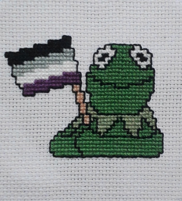 cross stitch of kermit the frog holding the asexual flag, on white aida.