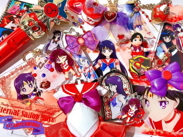 Photo of a spread of assorted merchandise, all focused on the character Sailor Mars from the franchise Pretty Guardian Sailor Moon. Her character design features long purple-black hair and purple eyes, with a red and purple costume inspired by Japanese sailor-style school uniforms. Merchandise includes various keychains featuring new and recycled key art of her character, a birthday cake-shaped charm with her image colours, a keychain styled like a little Kewpie doll wearing her costume, a tube of lip balm designed after her magical red transformation wand, a plastic French macaron with her colour scheme, and a plush stuffed keychain based just on her costume.