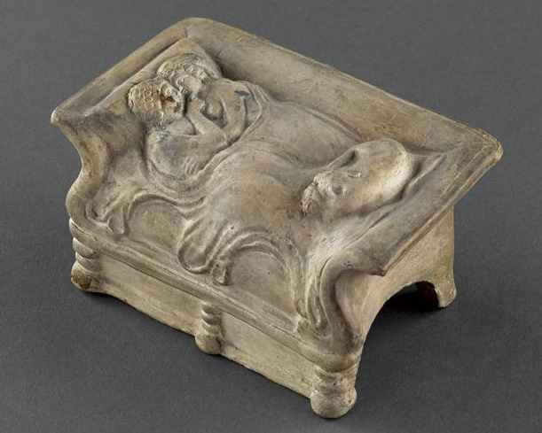 A terracotta model of two people embracing in bed, nude but under a blanket. One holds the other's chin. At the foot of the bed, a dog is curled up asleep.