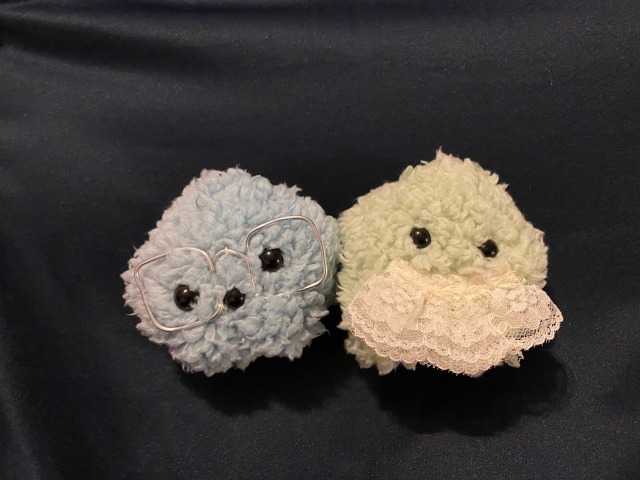 Two small star shaped plushes on a black background. the one on the left is blue and has glasses made of wire. the one on the right has a mustache made of lace trim.