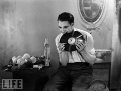 A man chews up a gramophone record. Beside him is a table with light bulbs about to be eaten, as well.