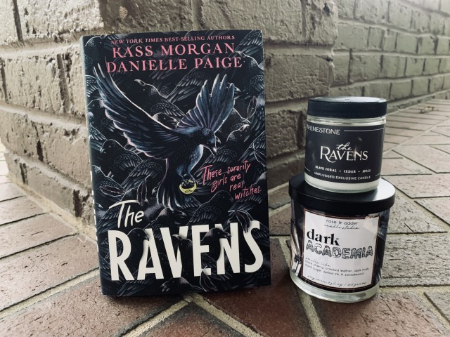 Shown is THE RAVENS (with cover art depicting ravens) beside two candles, one marked "Dark Academia" and one marked "The Ravens," against a brick backdrop. Photo by AHS.