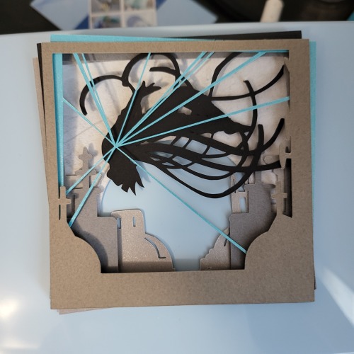 layers of paper in shades of grays, black, and bright blue stacked haphazardly together. They depict the silhouette of a girl in a cloak of long ribbons floating around her as she back flips over gray roof tops. Thin blue lines radiate from the center of her chest out to the edge of the frame.