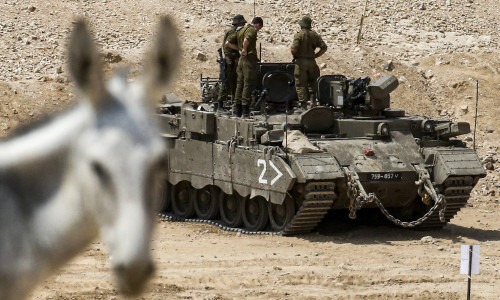 A donkey looks on as behind it Israeli soldiers stand aboard an IFVs, at a military training camp ne