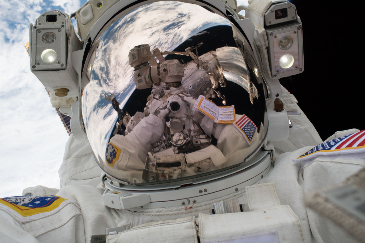 Astronaut Mark Vande Hei takes a selfie in front of Earth during the first spacewalk of 2018. His suit is white, the reflective helmet silver, and Earth is blue with white clouds. Credit: NASA