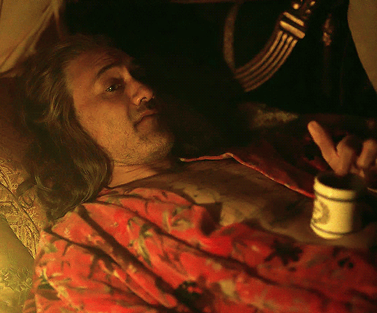 Gif of Edward Teach from Our Flag Means Death. He is laying on his back in a blanket fort and the camera captures him from the waist up. His head is propped up by pillows and he is lit by candlelight. He is wearing a pink robe with an ornate pattern, and the robe is open, revealing his bare chest. On his stomach rests a jar of marmalade, which Ed dips his index finger into before bringing his finger to his mouth, sucking the marmalade off his finger.