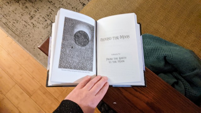 A handbound copy of Jules Verne's From the Earth to the Moon and Around the Moon, opened to the title page for Around the Moon. It has an illustration of a small vessel orbiting the moon. 