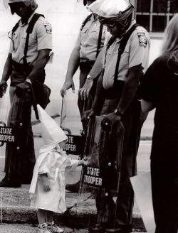 plainwhitebread:  brain-food: Here is a Georgia State Trooper in riot gear at a KKK protest in a north Georgia city back in the 80s. The Trooper is black. Standing in front of him and touching his shield is a curious little boy dressed in a Klan hood