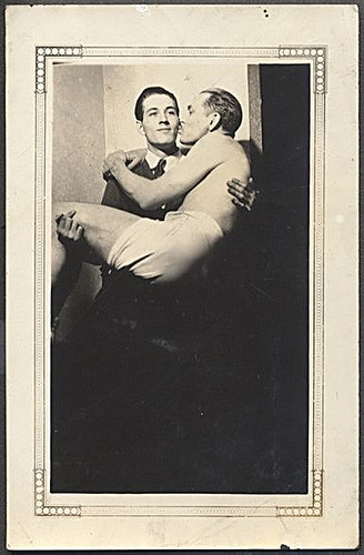 A vintage photograph of a man holding another. The man getting held is shirtless and in his underwear.