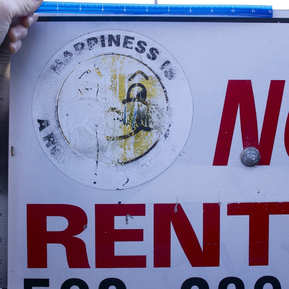 Apartment rental sign with graffiti and significant weather damage, smiley face logo, “HAPPINESS IS A RENT GUARANTEE”. N Fessenden and N Wall.