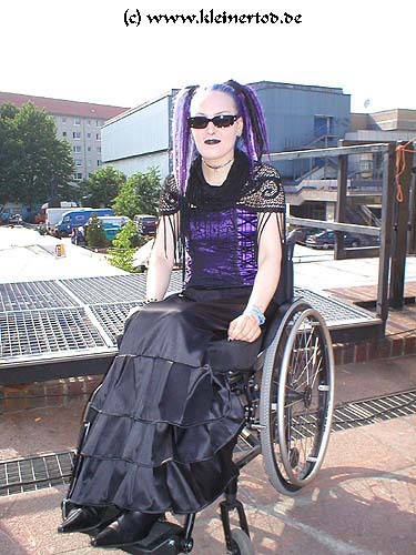 photo of a person in a wheelchair wearing a nice corset + skirt ensemble, along with some pointy boots and purple dreadfalls