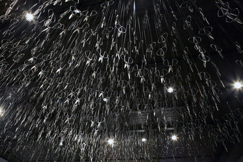  Beili Liu - The Mending Project (2011) “…Hundreds of Chinese scissors suspended from the ceiling in a shimmery cloud. The piece involved the artist sitting at a small black table, hand-mending patches of fabric together which visitors were encouraged