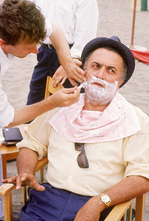 avagardner:Federico Fellini receives a shave while sitting on his director’s chair in the sand, 1965