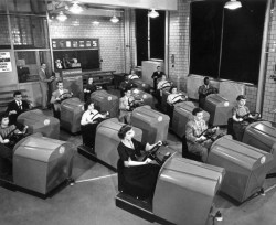 Students at Brooklyn High School in New York learn to handle the controls of a car and experience simulated traffic conditions flashed onto a screen by means of projected film, using the Aetna Drivotrainer.