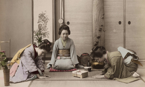 Hand-colored photo of two young women greeting another woman, presenting her with a gift box tied wi